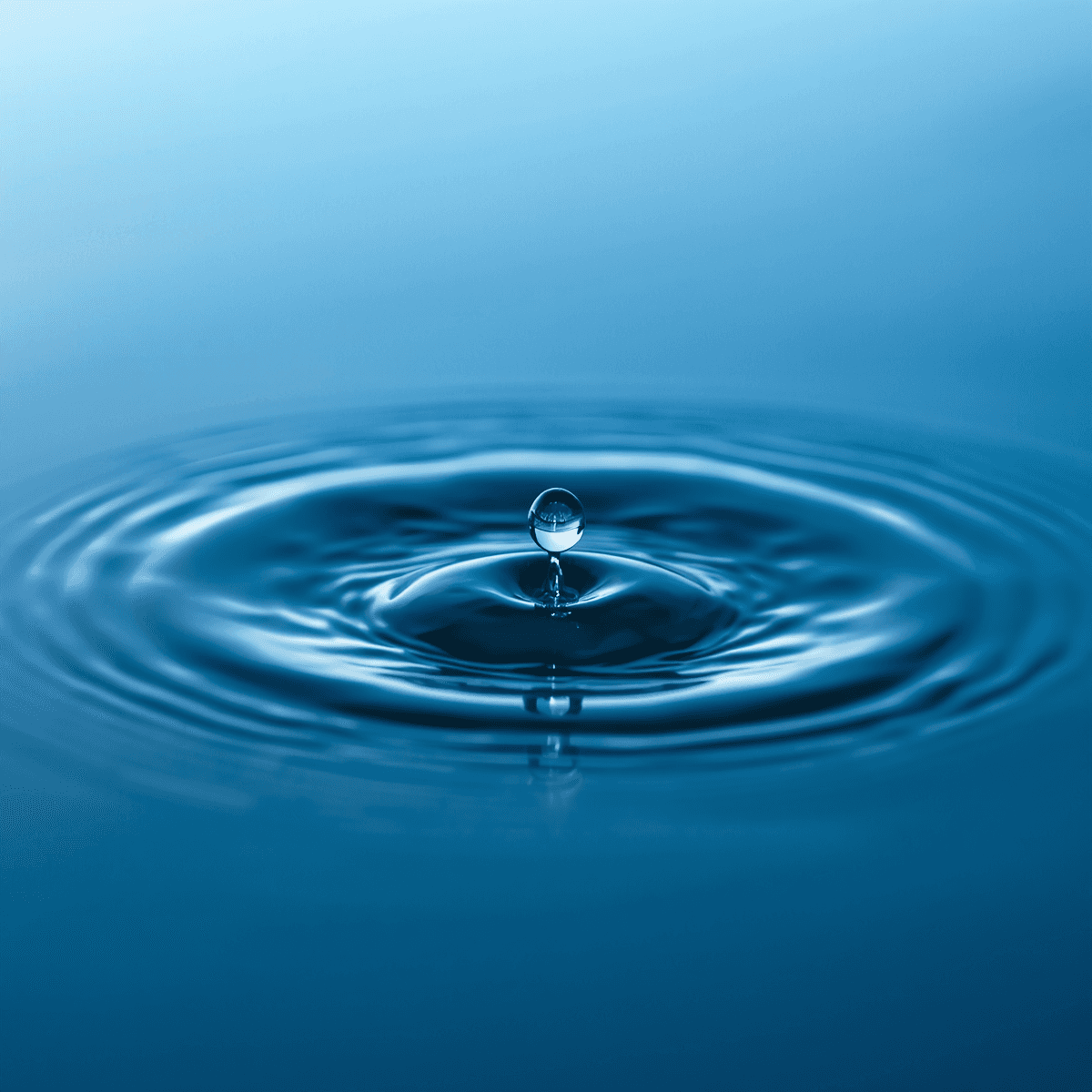 Single water droplet suspended above blue water, creating concentric ripples on the smooth surface.