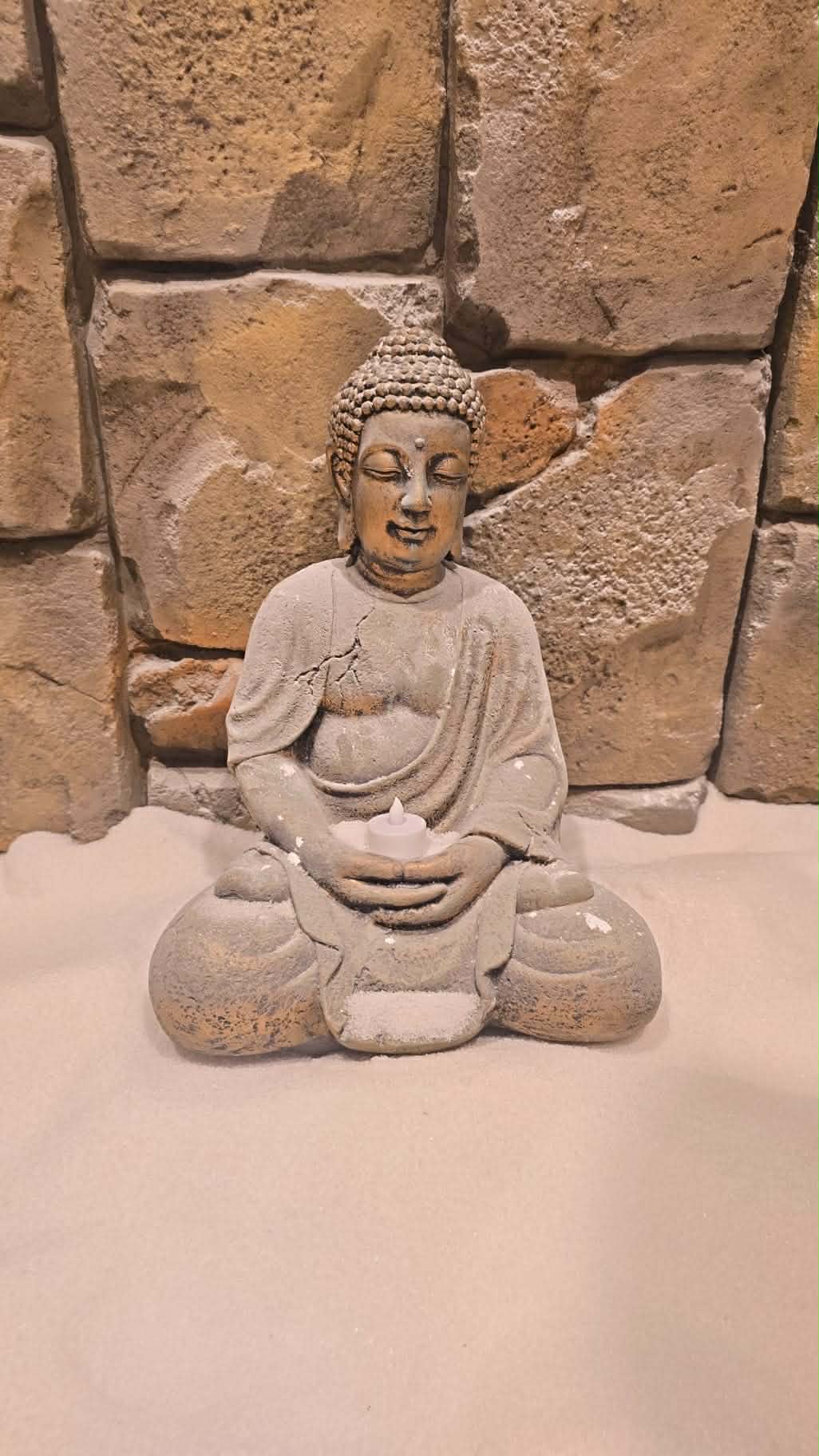 Weathered Buddha statue in meditation pose holding a candle on sand before a stone wall.